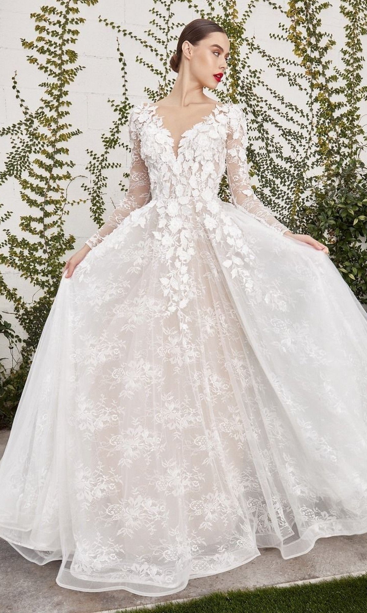 A woman stands outdoors against a wall covered in green vines, wearing the Andrea and Leo A1067WC long formal dress in off white/nude, featuring long sleeves and exquisite floral lace appliques.